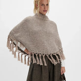 Model wearing a neutral-toned mock neck knit poncho with tassel trim, styled over a long-sleeve top against a clean studio background.