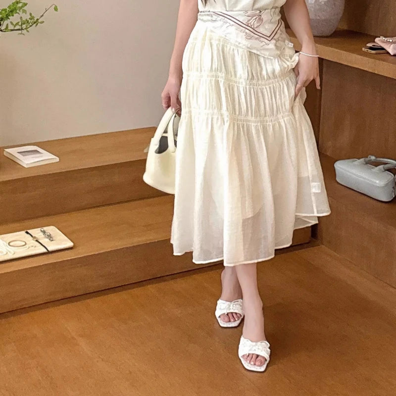 Woman in a beige dress holding a white handbag in a room with wooden floors and shelves.
