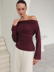 Model wearing an off-shoulder long sleeve top with a tie-up detail, styled with wide-leg trousers in a minimalist interior setting.
