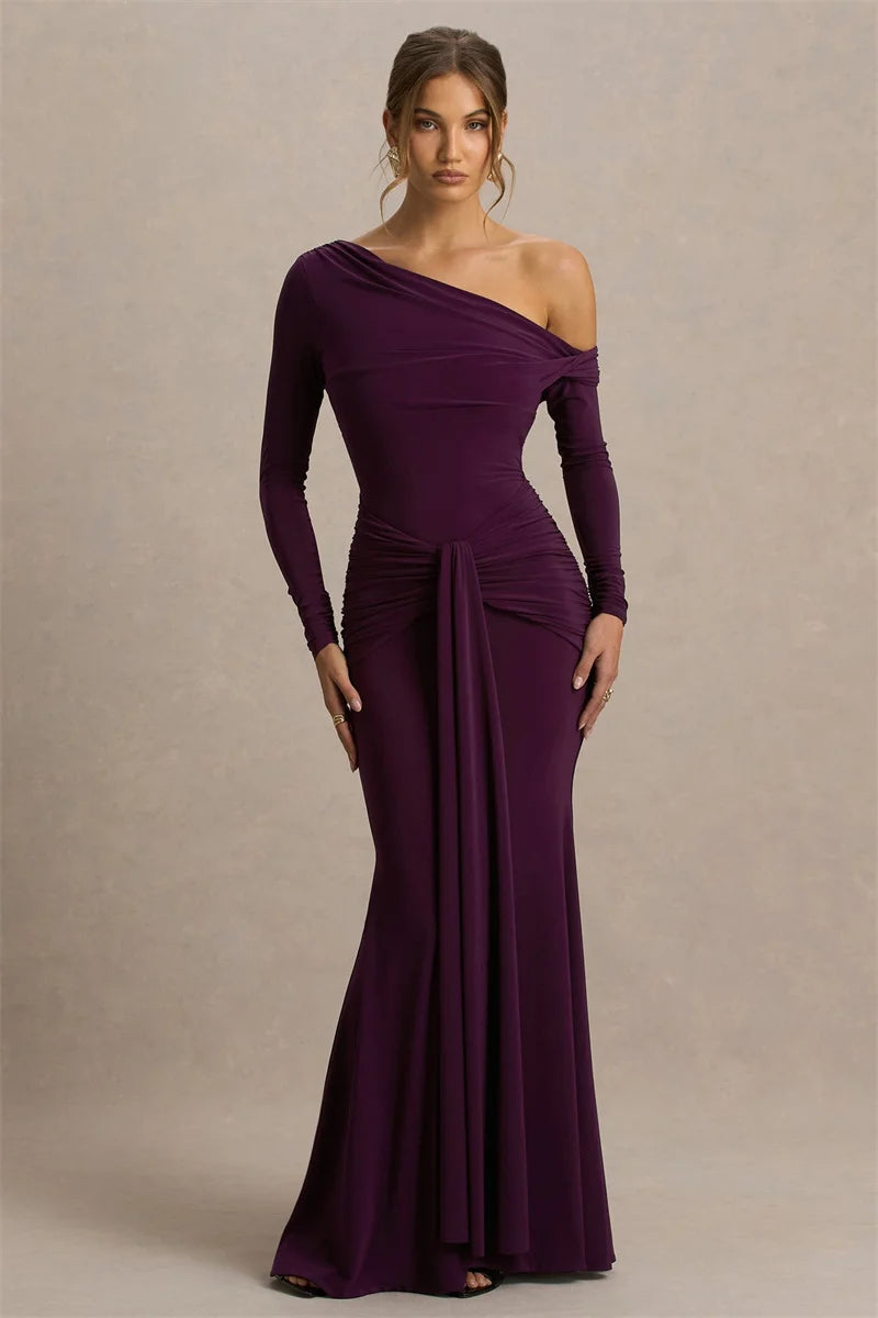 Model wearing a deep purple one-shoulder maxi gown with a single long sleeve, standing in studio lighting with a sculpted, elegant silhouette.