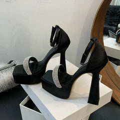 Black high-heeled shoes with rhinestone straps on a white platform, placed on a white surface.