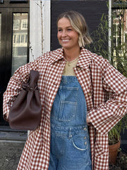 Woman wearing a red and white checkered coat over denim overalls, holding a brown bag.