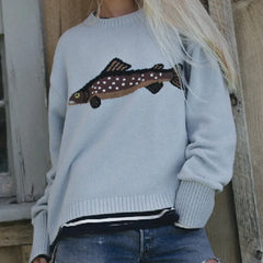Light blue sweater with fish design worn by a person, standing against a wooden wall.