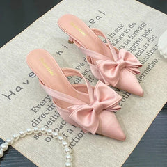 Pointed-toe bow mules in pink are styled for elegant summer occasions on a textured surface