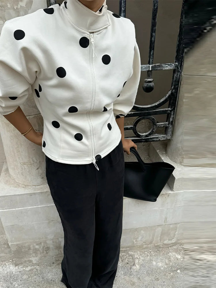 Person wearing a white jacket with black polka dots and black pants, holding a black handbag.