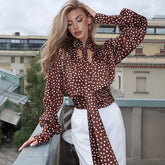 Woman wearing brown polka dot satin lantern sleeve blouse styled with white trousers on city balcony.