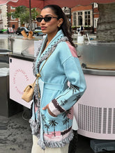 Street-style shot of a woman in a light blue printed long cardigan with tassel hem, sunglasses, and a crossbody bag near a kiosk.