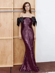 Model wearing a purple sequin sheath maxi with black off-shoulder bows, posed against warm wooden panels and a white wall.