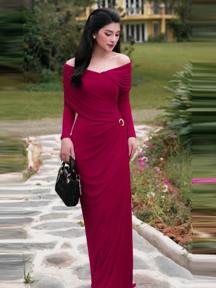 Woman in a red asymmetrical off shoulder long gown with metal pin styled for parties standing on a stone path with greenery in the background