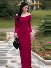 Woman in a red asymmetrical off shoulder long gown with metal pin styled for parties standing on a stone path with greenery in the background