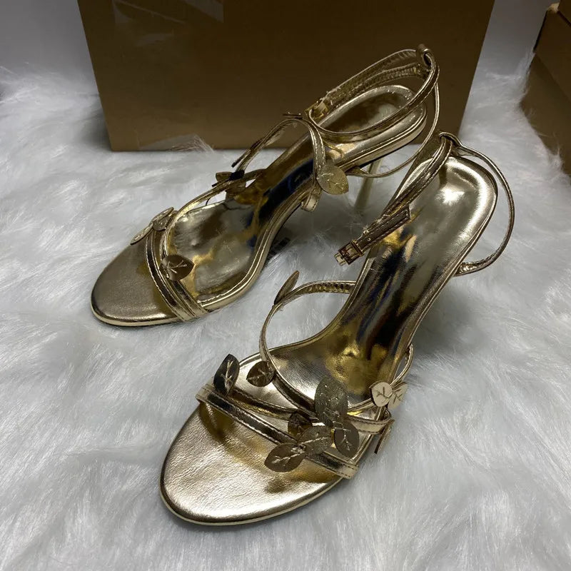 Gold high-heeled sandals on a white fur surface with a brown box in the background.