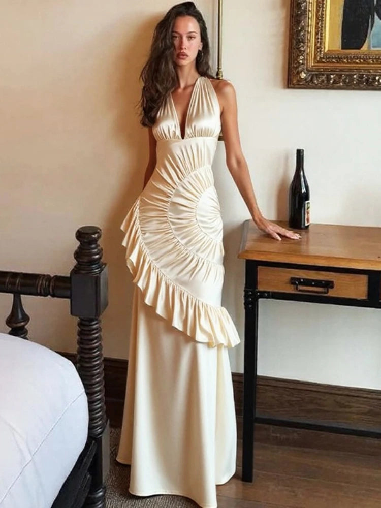 Editorial view of a satin ruched neck-mounted maxi dress styled in a bedroom setting with floor-length pleated sweep.