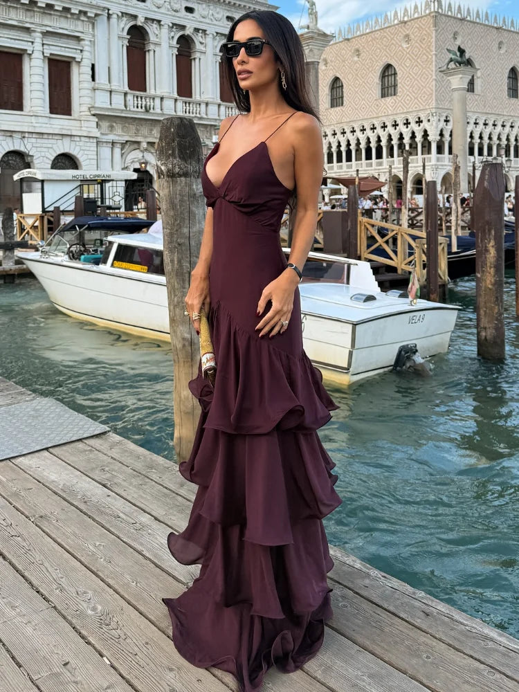 Woman in a burgundy satin cami maxi dress standing on a waterfront dock, featuring a V-neckline and tiered ruffle skirt.