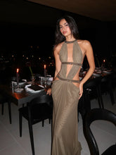 Woman in a brown-gold sequin halter maxi dress standing in a dimly lit dining space, styled with minimal accessories for evening elegance.