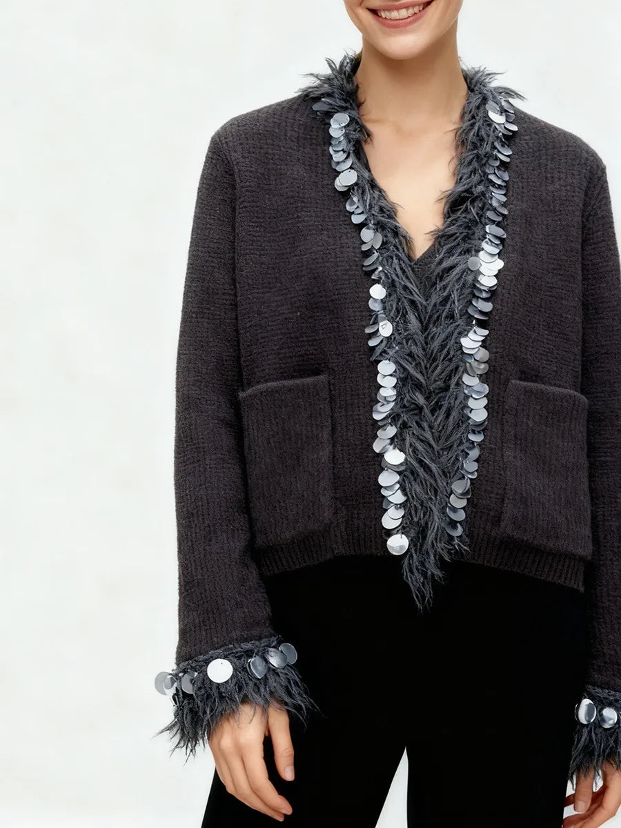 Relaxed knit jacket with sequin and tassel trim styled casually over dark trousers in a minimalist studio setting.