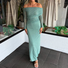 Person wearing a close-up of a teal asymmetric neckline, sheer woven blouse with folded detailing in an indoor setting with plants and clothing in the background.