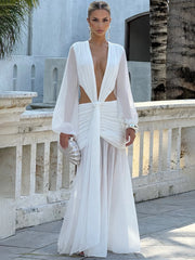Woman in a sheer-sleeved evening dress styled for resort parties standing on a balcony with architectural elements in the background.