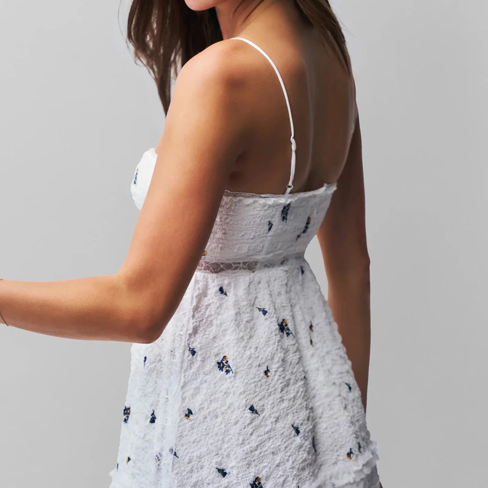 A woman wearing a white summer mini dress with black flower accents and adjustable straps on a grey background