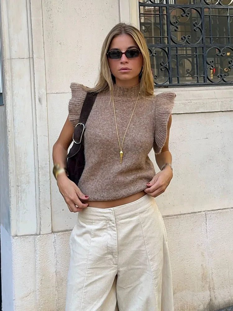 Model wearing a brown sleeveless knit top with ruffled shoulders, styled with relaxed trousers against a textured stone wall.