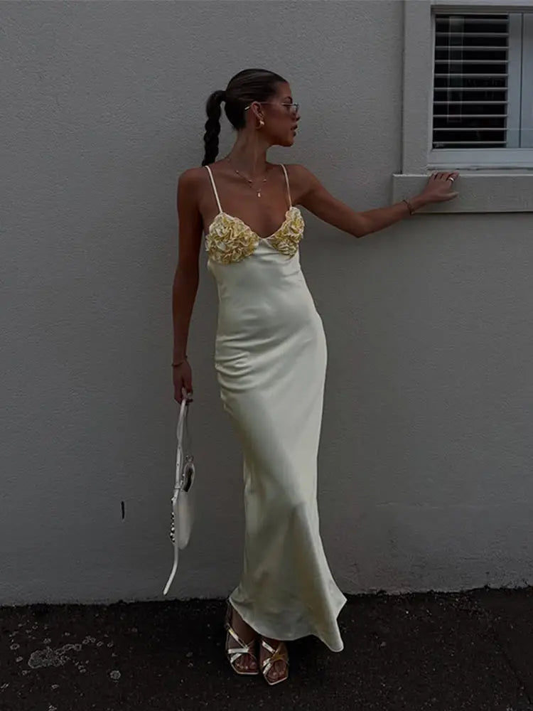 Woman in a white evening gown with floral details against a plain wall.