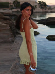 A woman in a backless green knitted mini dress with tassel hemline standing by a body of water with rocks and a building in the background.
