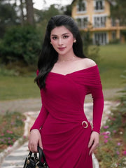 Woman in a slim ankle length evening dress in red for formal celebrations, standing outdoors with greenery and a building in the background