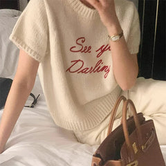 Person wearing a cream sweater with 'See ya Darling' text, sitting on a bed with a brown handbag.