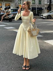 Woman in a square-neck fall dress with long sleeves and pleated skirt holding a wicker bag and ice cream cone on a city street.