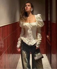 Studio shot of a woman wearing an ivory square neck corset blouse with puff sleeves, styled with black leather-look pants on a grey backdrop.