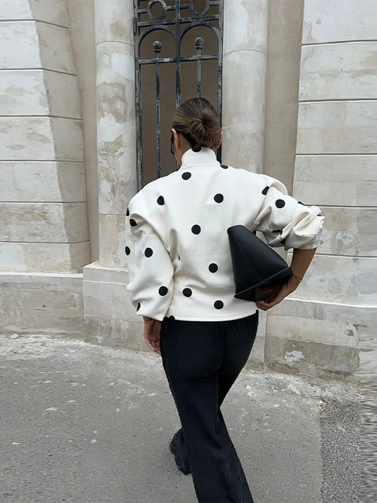 Person wearing a white polka dot jacket and black pants, holding a black clutch, standing against a stone building.