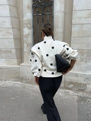 Person wearing a white polka dot jacket and black pants, holding a black clutch, standing against a stone building.