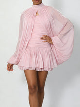 Model wearing a pale pink strapless ruched mini dress with a sheer cape overlay, posed with one hand on hip in studio light.