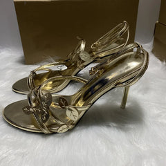 Gold high-heeled sandals on a white fur surface with a brown box in the background.