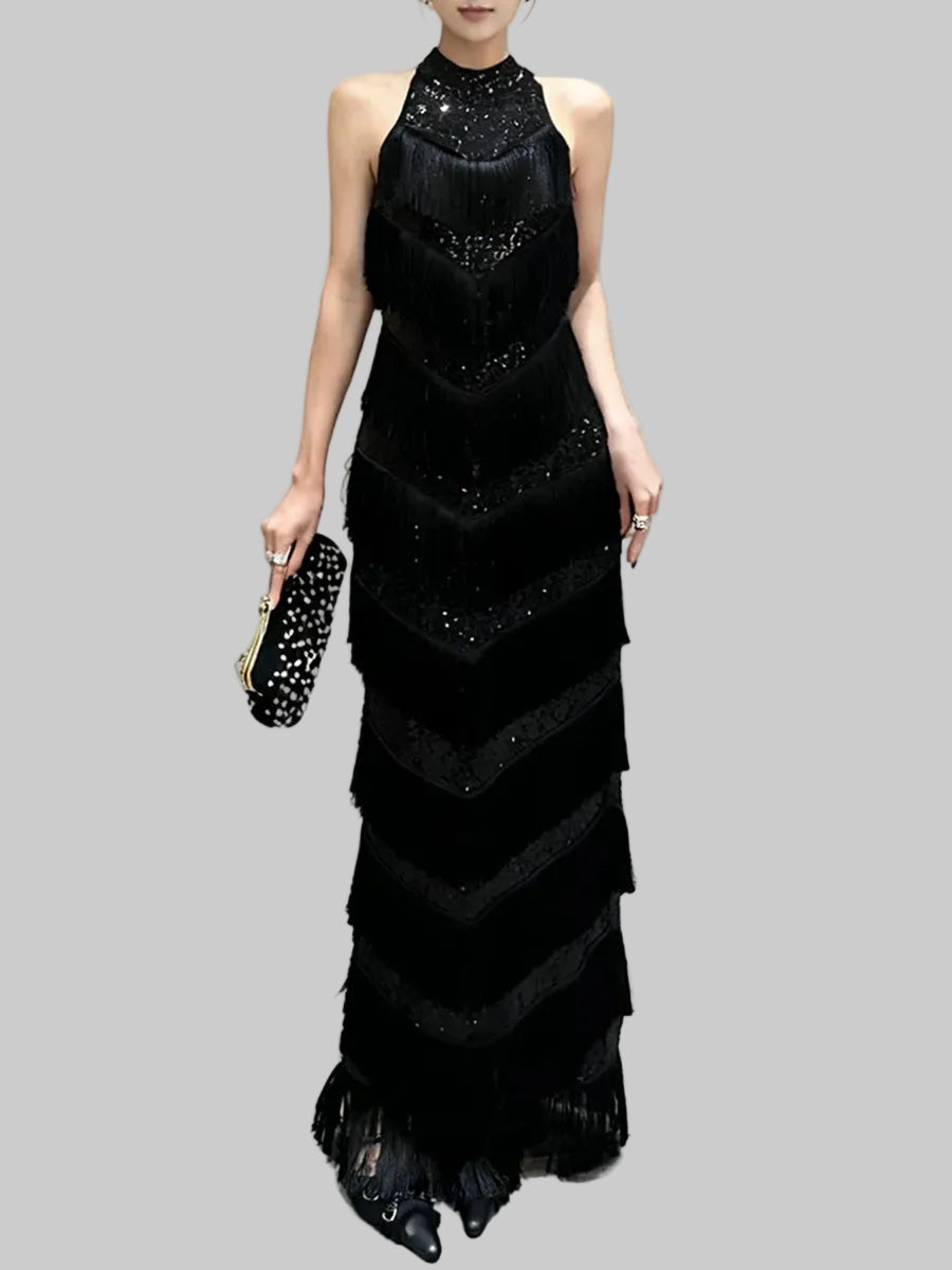 Full-length studio image of a black sleeveless halter maxi dress with tiered tassel bands creating a dramatic, elongated silhouette.