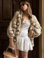 Woman wearing a beige cardigan with black patterns and a white dress, standing in a dimly lit room.