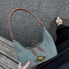 Blue denim handbag with brown leather strap held by a person on a textured surface.