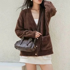 Person wearing a brown cardigan holding a vintage-inspired coffee brown satchel with dual handles and a zipper closure against a plain background