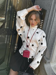Woman wearing a white jacket with black polka dots, holding a red handbag, standing against a building entrance.
