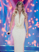 Model wearing a white halter maxi dress with a draped neckline and delicate bead detailing, photographed under colorful stage-style lights.