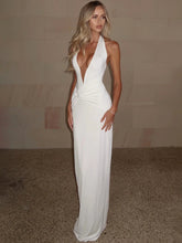 Model in a white halter maxi dress with a deep V neckline, standing against a neutral wall with a clean, elongated silhouette.