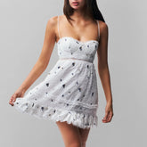 A woman wearing a white lace mini dress with black floral embroidery and ruffled hem on a grey background.