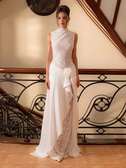 Woman in a white lace patchwork maxi dress standing by an ornate staircase railing, showing a sleek sleeveless bodice and flowing skirt.