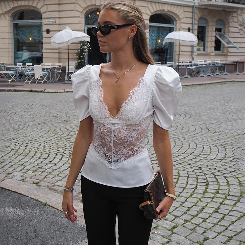 Street-style look featuring a white puff-sleeve plunge top with lace panel, paired with black pants and sunglasses on cobblestones.