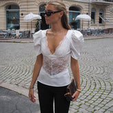 Street-style look featuring a white puff-sleeve plunge top with lace panel, paired with black pants and sunglasses on cobblestones.
