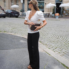 Outfit view with the white lace-front top styled with tailored black pants and a clutch, framed by café seating in the background.