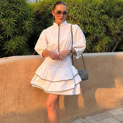 Woman in a white puff sleeve mini shirt dress with stand collar  standing outdoors with greenery in the background
