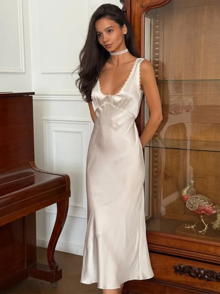 Woman wearing a white satin slip dress with lace V-neckline styled for summer parties standing in a room with wooden furniture.