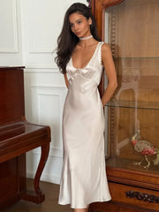 Woman wearing a white satin slip dress with lace V-neckline styled for summer parties standing in a room with wooden furniture.