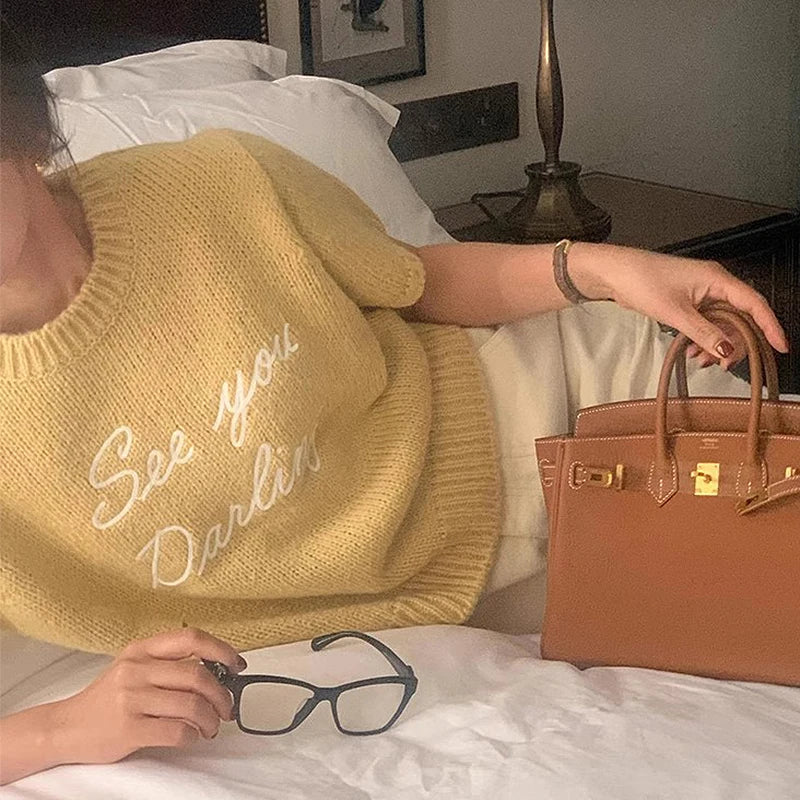 Person wearing a yellow sweater with text, holding glasses and a brown handbag on a bed.