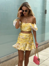 Woman wearing yellow floral two-piece skirt set with puff sleeves carrying a pink hand bag.
